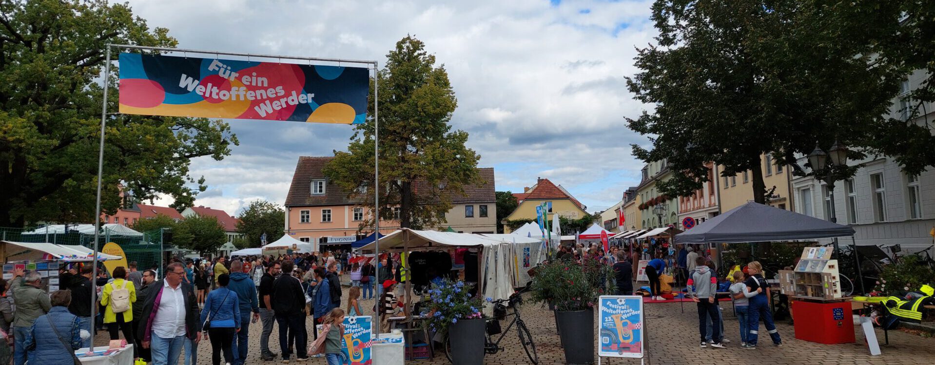 Foto des Marktplatzes von Werder am Tag des Festival Weltoffenes Werder. Viele Menschen sind auf dem Platz, dort stehen Marktstände und ein großes buntes Banner mit dem Schriftzug "Für ein weltoffenes Werder" sind zu sehen.